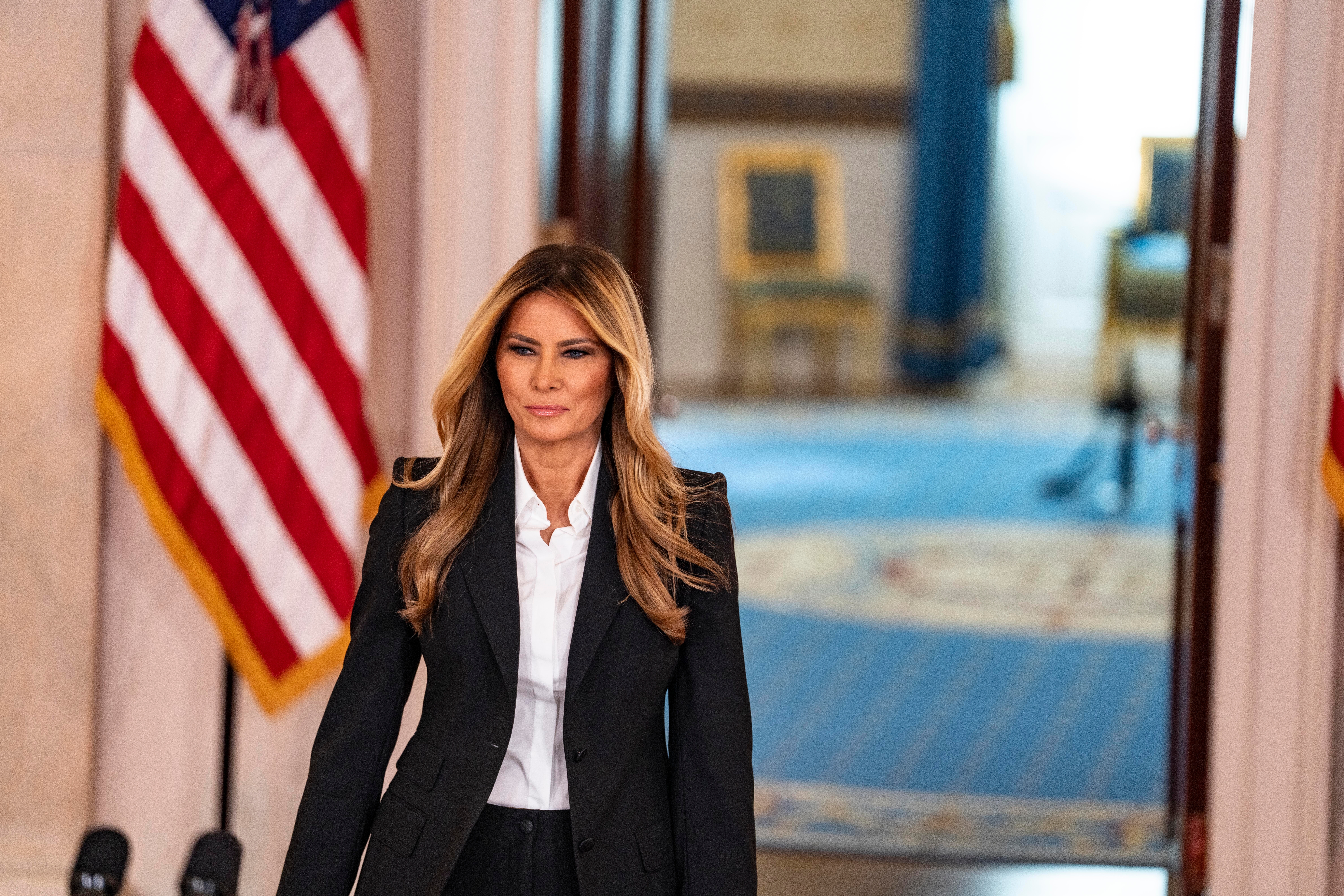 Mujer vestida con traje negro y camisa blanca camina por un pasillo institucional, con banderas de Estados Unidos al fondo, en un entorno oficial.