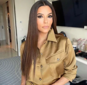 Mujer con cabello lacio castaño oscuro posa frente a cámara en un interior moderno, vestida con una camisa satinada color dorado de manga larga y maquillaje sofisticado en tonos neutros.