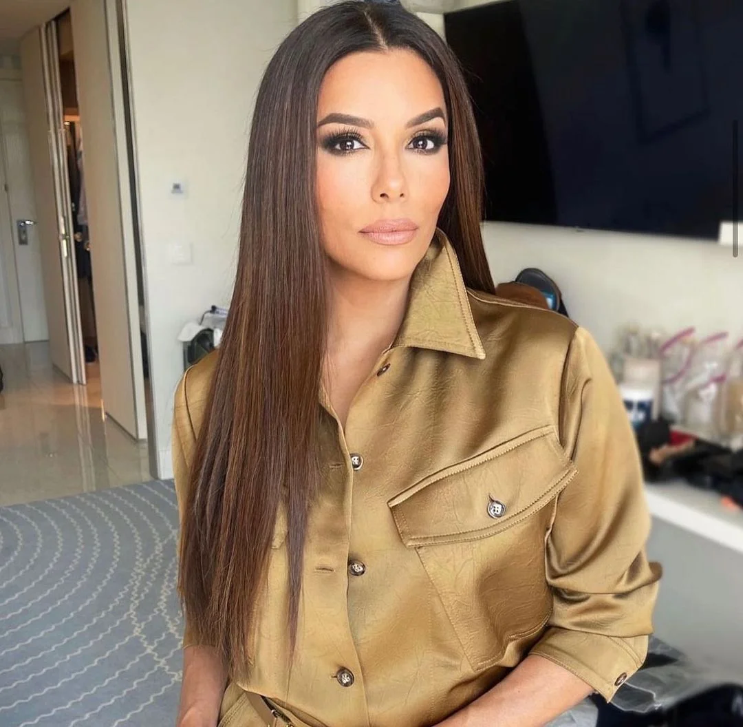 Mujer con cabello lacio castaño oscuro posa frente a cámara en un interior moderno, vestida con una camisa satinada color dorado de manga larga y maquillaje sofisticado en tonos neutros.
