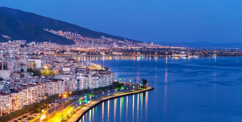 Vista nocturna de una ciudad costera iluminada, con edificios junto al mar y una avenida curvada bordeando la bahía.
