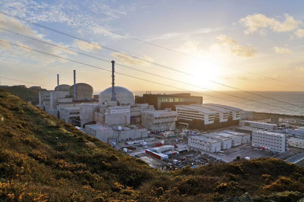 Vista panorámica de una central nuclear junto a la costa, con reactores, edificios industriales y el mar al fondo durante el atardecer.