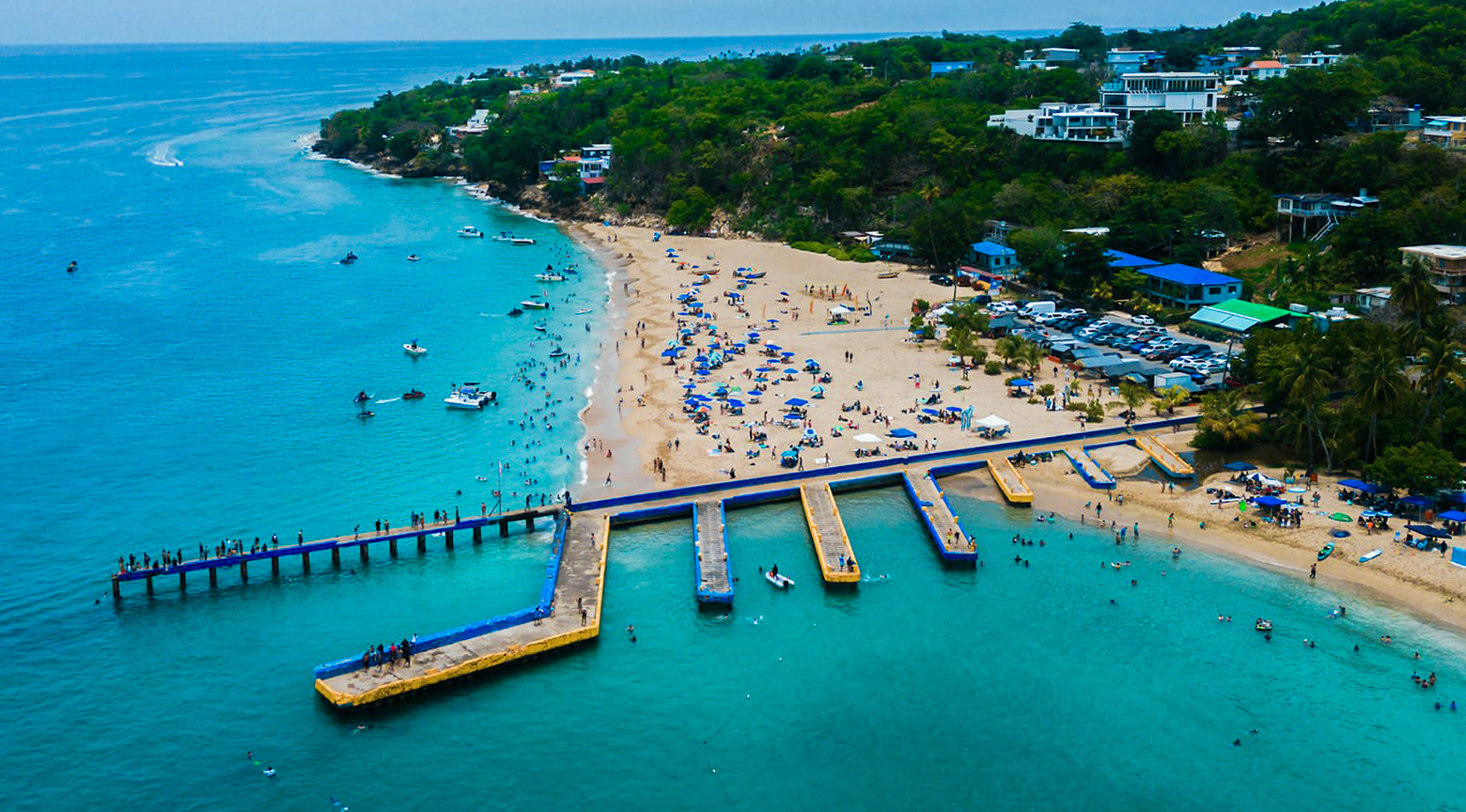 Vista aérea de la playa de Aguadilla con muelles, embarcaciones y bañistas sobre aguas turquesa.