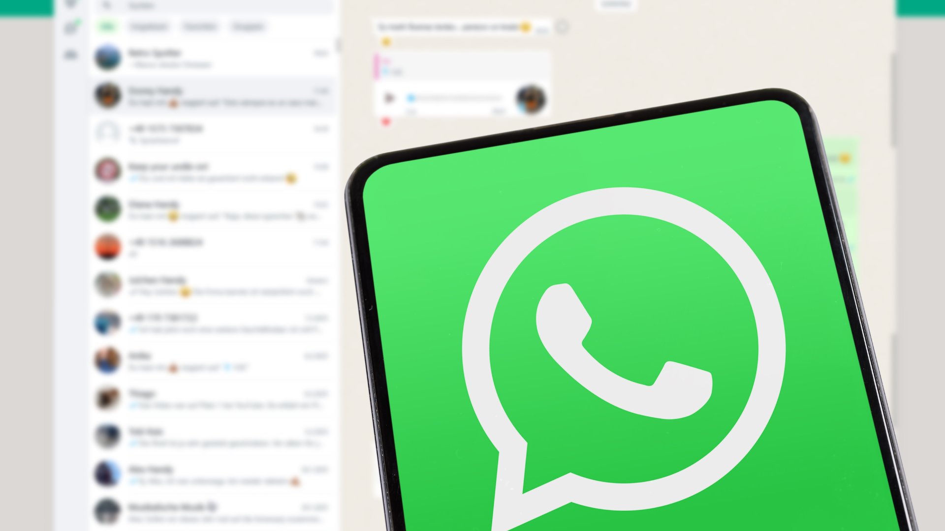 Pantalla de un teléfono móvil con el logo de WhatsApp en primer plano y una lista de chats desenfocados al fondo.