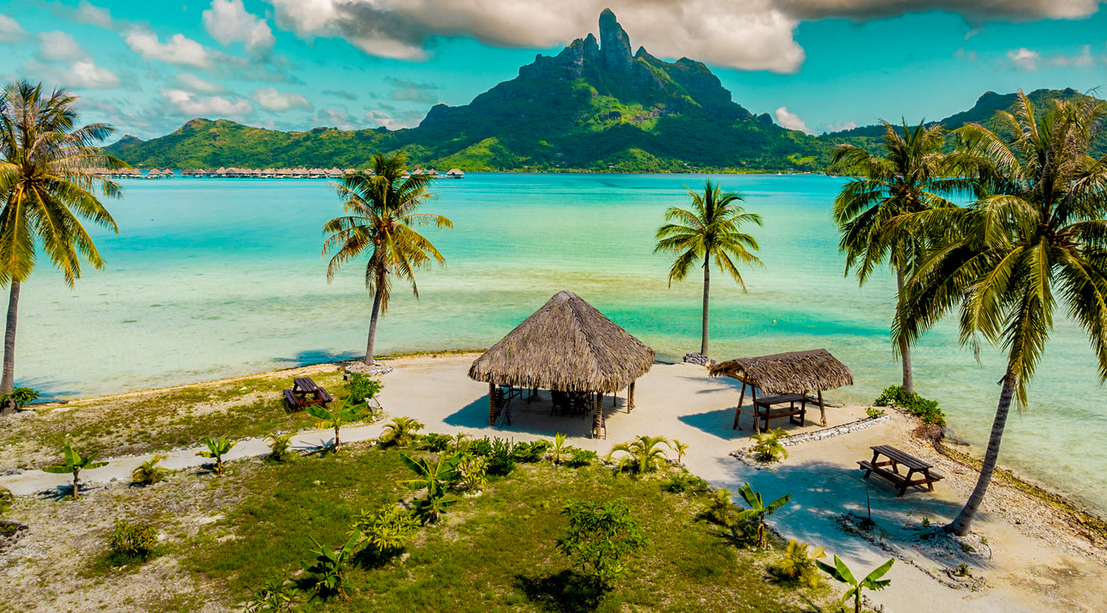 Playa de Bora Bora con palmeras, cabañas de paja y una laguna turquesa frente a montañas verdes.