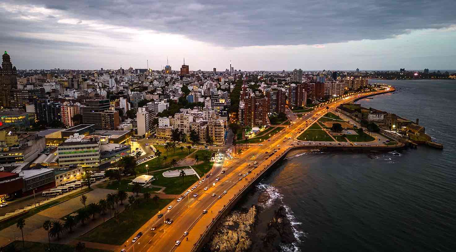 Vista aérea de una ciudad costera al atardecer, con una avenida iluminada junto al mar y edificios urbanos al fondo.