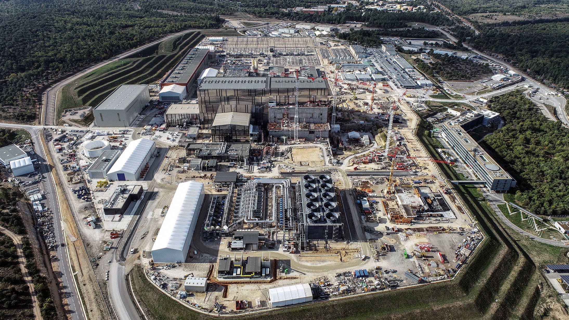 Vista aérea del complejo ITER en construcción, con edificios industriales, grúas y estructuras tecnológicas rodeadas de áreas verdes.