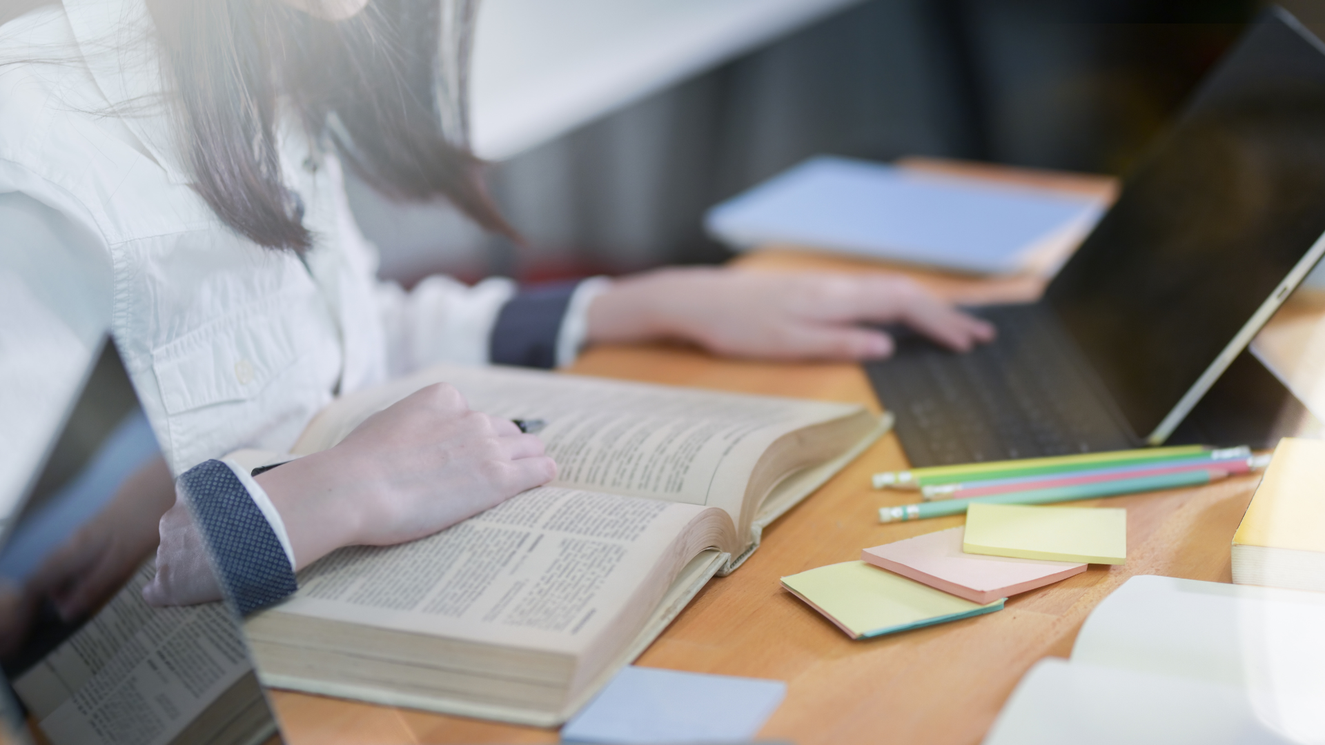 Persona estudiando en un escritorio con un libro abierto, una computadora portátil y materiales de estudio.