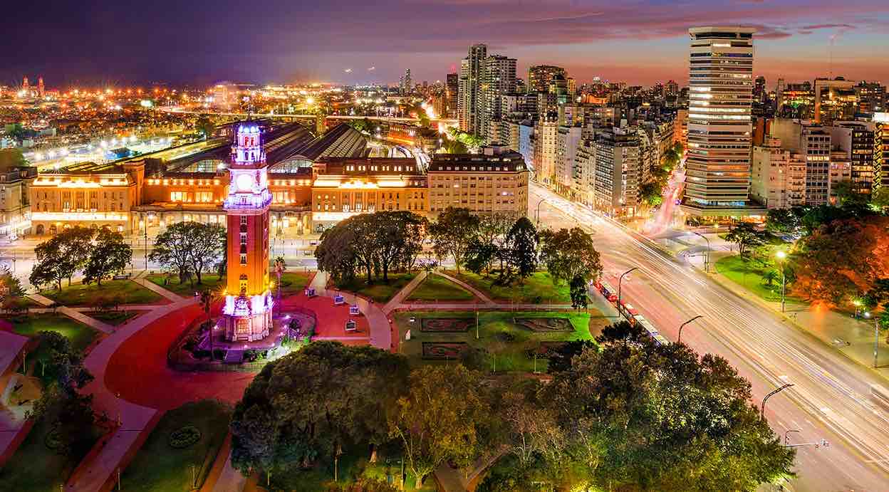 Vista aérea nocturna de Buenos Aires con avenidas iluminadas, edificios modernos y un monumento histórico en primer plano.