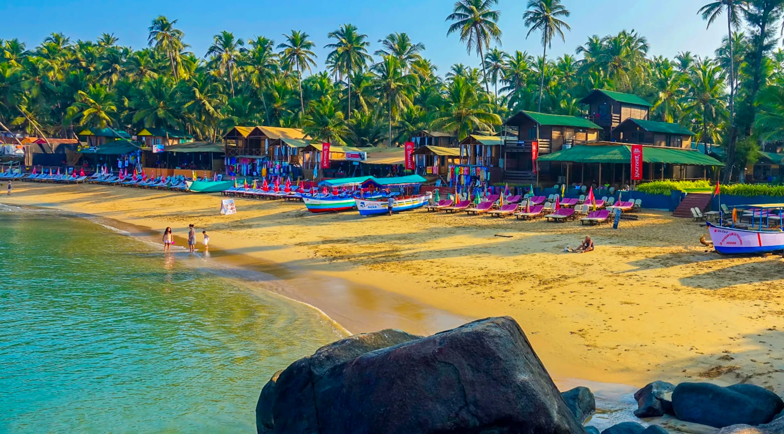 Playa de Palolem en Goa con palmeras, cabañas de madera, sombrillas de colores y botes frente al mar.