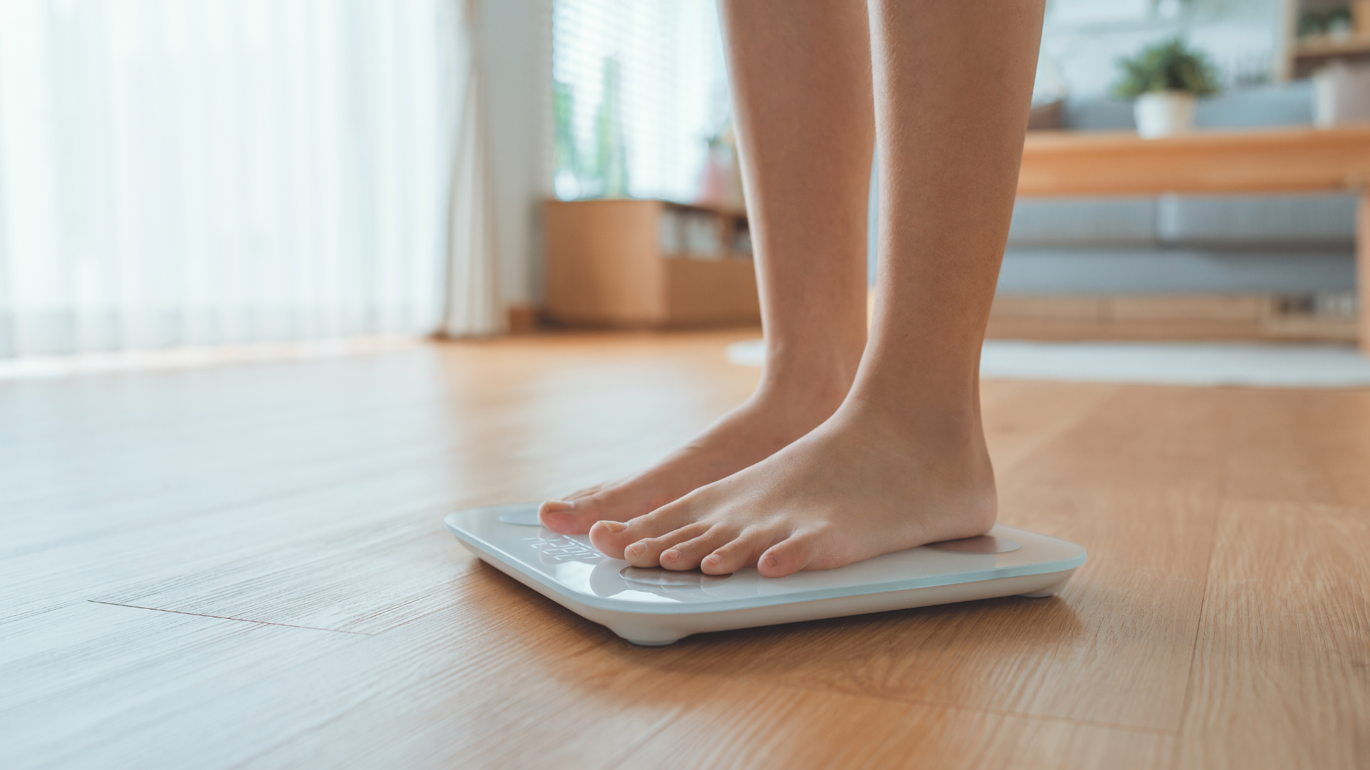 Persona de pie sobre una báscula digital en el interior de una vivienda, mostrando solo las piernas y los pies descalzos sobre un suelo de madera.