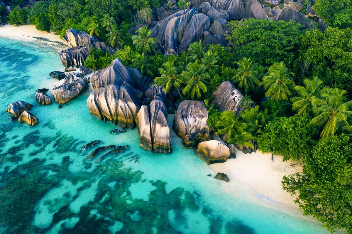 Vista aérea de una playa tropical con formaciones rocosas de granito, palmeras y aguas turquesas junto a una franja de arena blanca.