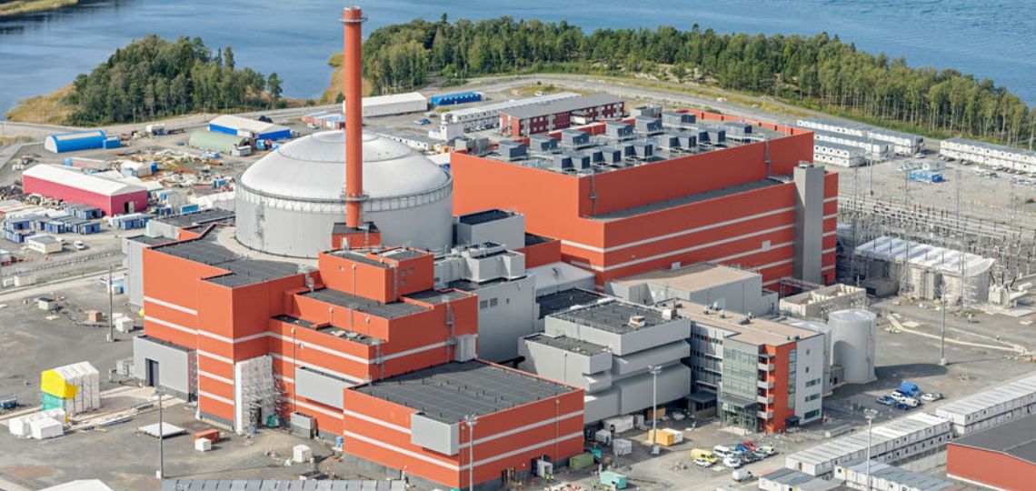 Planta nuclear con edificios industriales de gran escala, chimenea central y estructuras de seguridad, ubicada junto a un cuerpo de agua y rodeada de áreas verdes.