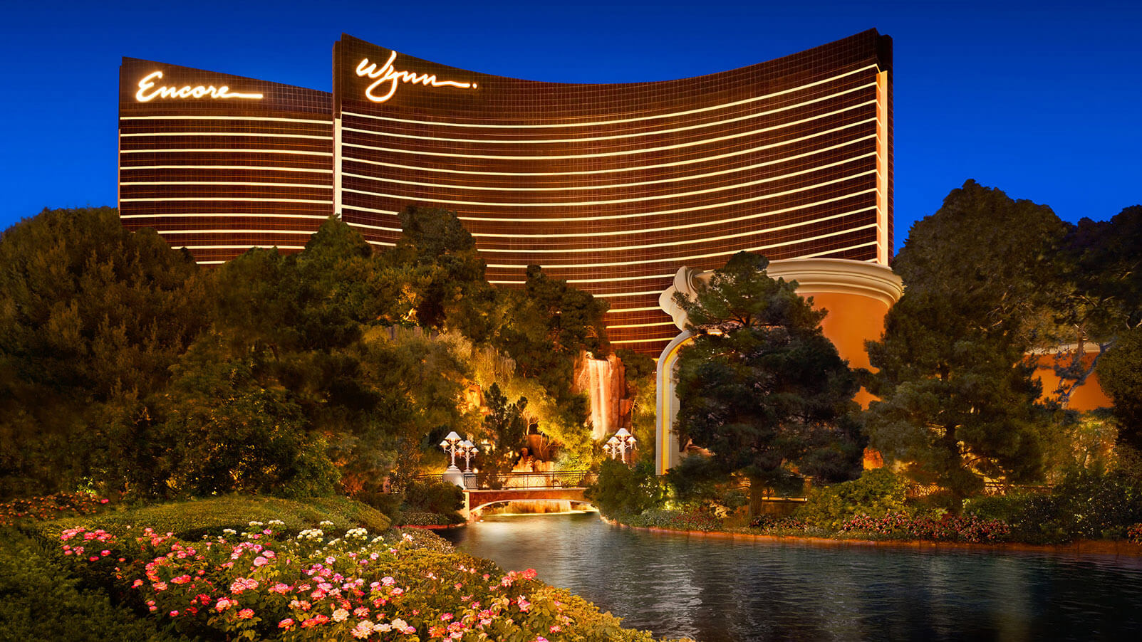 Vista nocturna del hotel Encore y Wynn Las Vegas iluminados en tonos dorados, con jardines, flores y un lago artificial en primer plano.