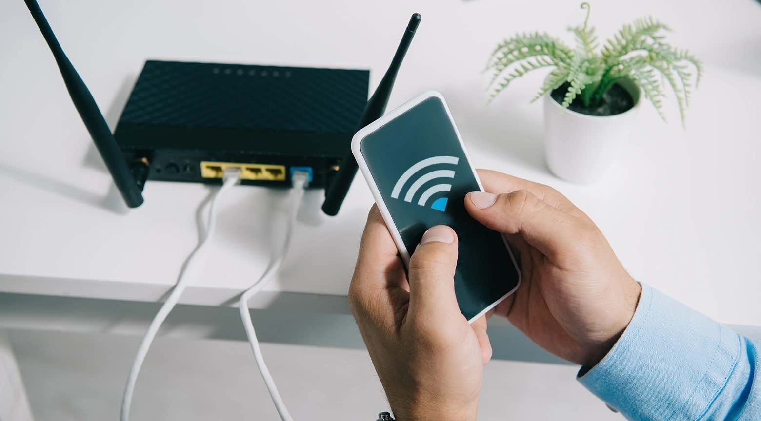 Persona sosteniendo un teléfono móvil frente a un router WiFi doméstico, con el ícono de señal inalámbrica en la pantalla y varios cables conectados al dispositivo.
