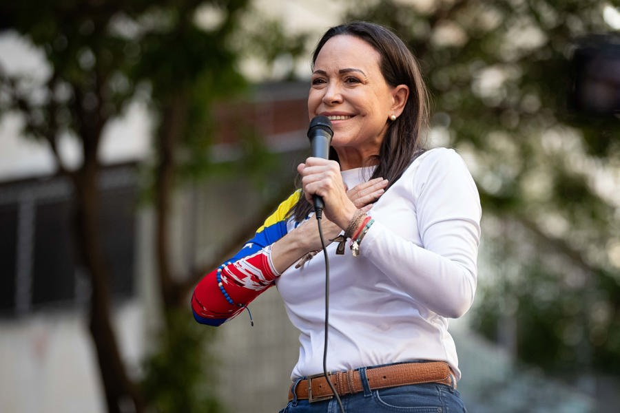 Mujer sosteniendo un micrófono durante un acto público, con la mano sobre el pecho y una banda con los colores de la bandera venezolana en el brazo.