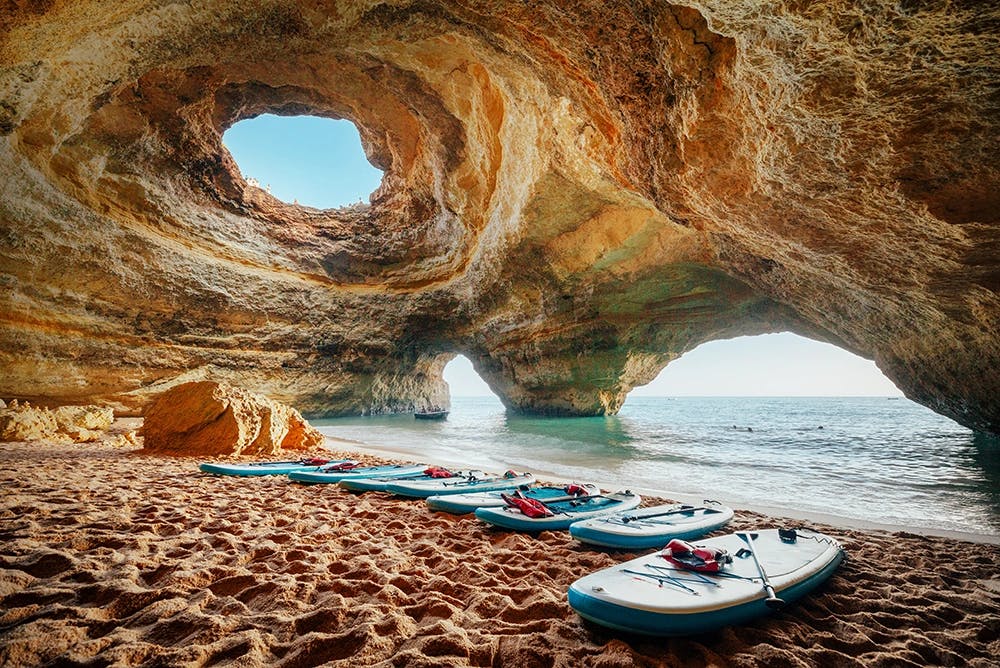 Tablas de paddle surf alineadas sobre la arena dentro de una cueva marina, con el mar y la luz entrando por las aberturas de roca.