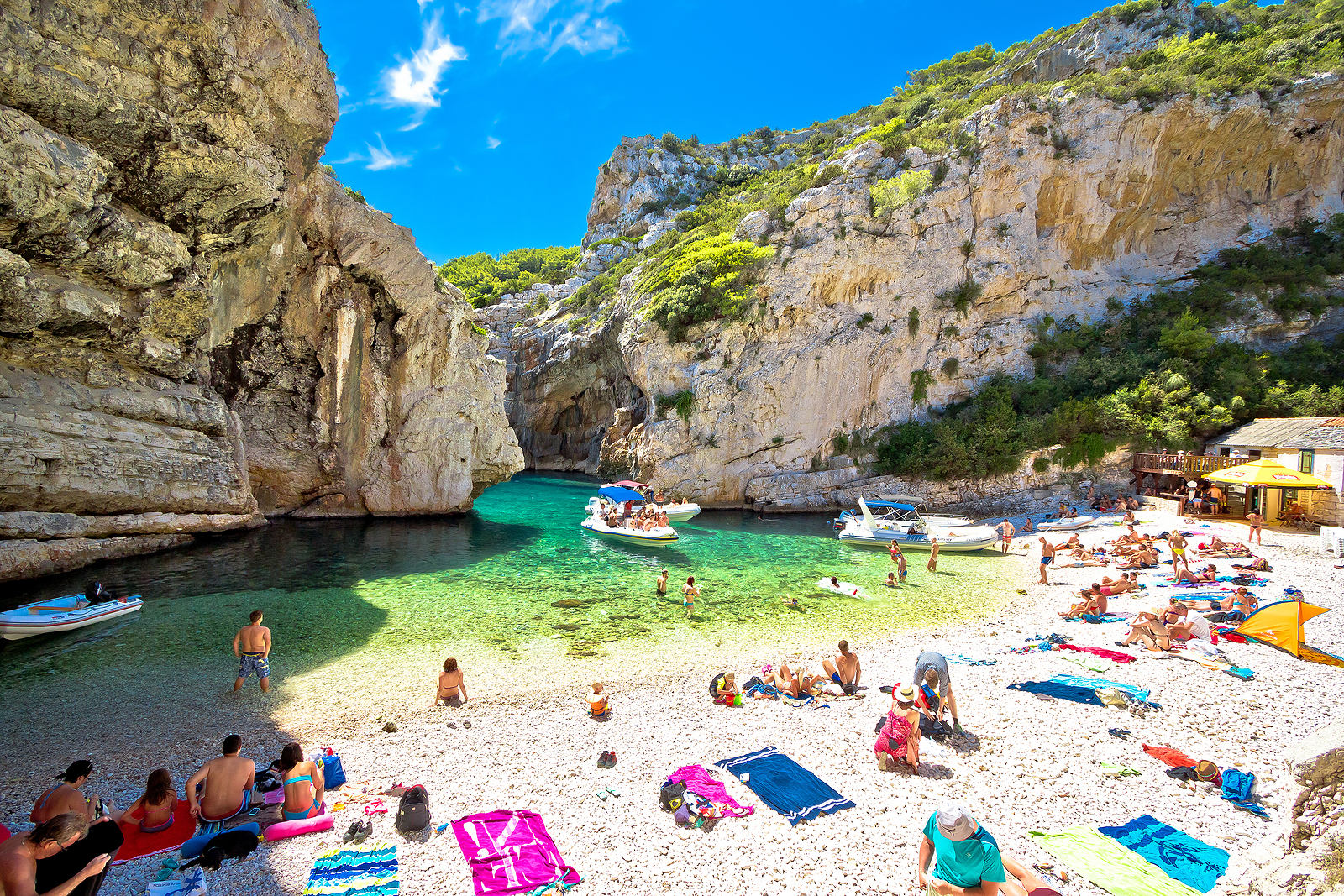Personas disfrutan de una cala de aguas cristalinas rodeada de acantilados rocosos, con embarcaciones ancladas cerca de la orilla.