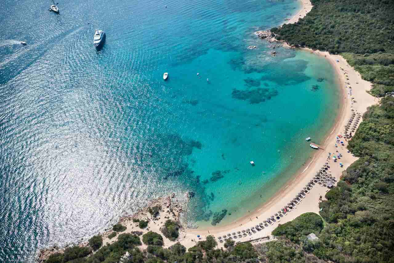 Vista aérea de una cala de aguas turquesas con playa en forma de media luna, embarcaciones y sombrillas alineadas en la orilla.