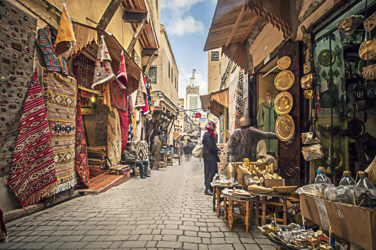 Calle estrecha de un zoco en Fez, con alfombras, telas y artesanías colgadas en los comercios y personas caminando entre los puestos.