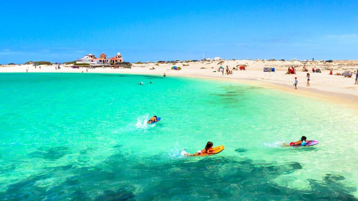 Personas disfrutan del mar turquesa en una playa de arena blanca, con dunas y edificaciones al fondo en un día soleado.