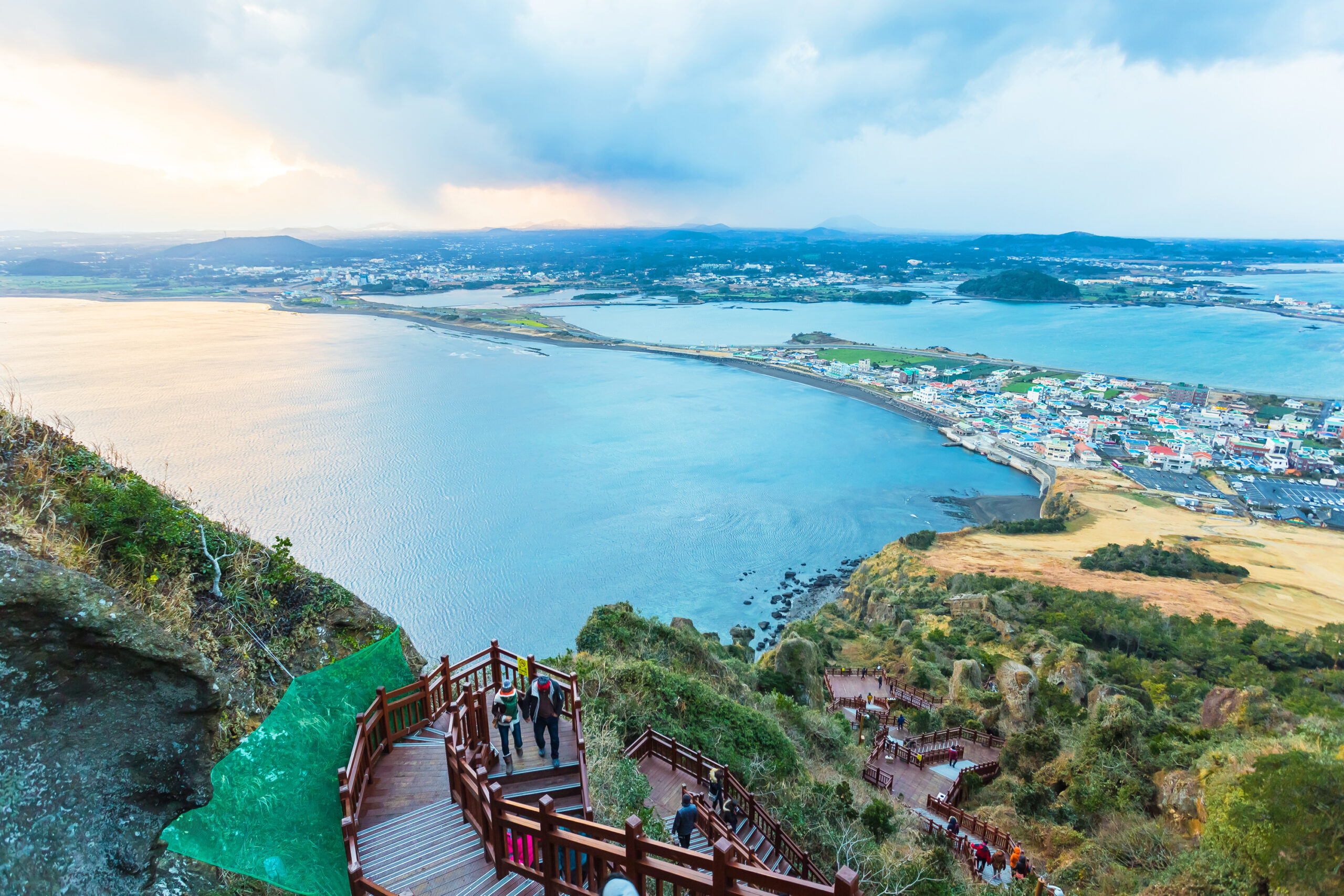 Vista panorámica de la costa de la isla de Jeju desde un mirador con escaleras de madera y visitantes recorriendo el sendero.