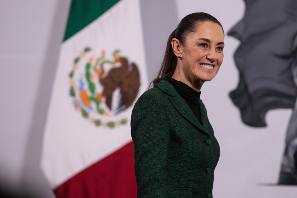 Mujer sonriente con traje verde camina frente a una bandera de México en un acto oficial.