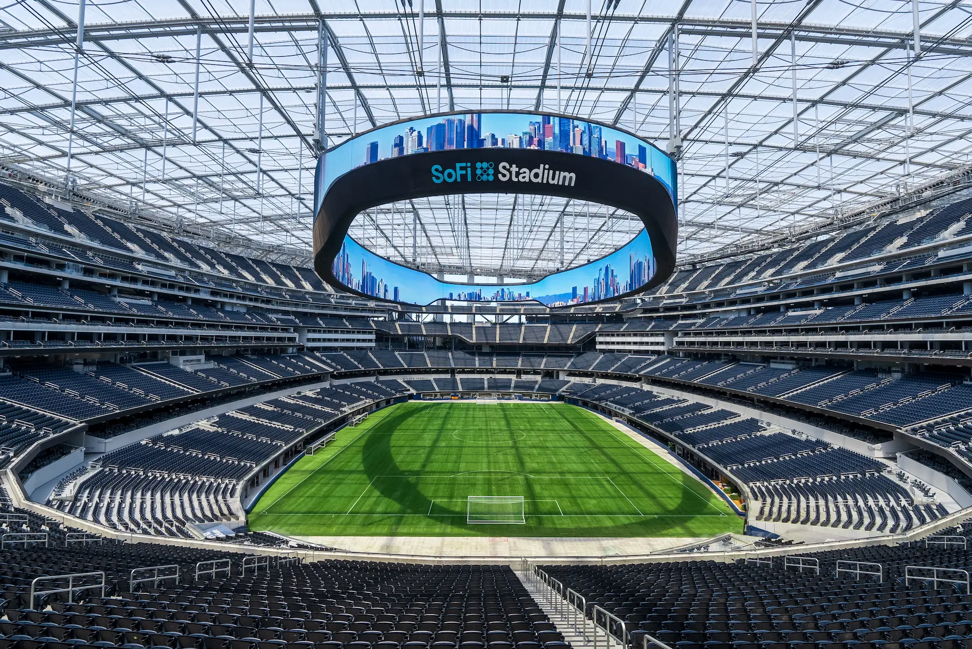 Vista interior del SoFi Stadium con el campo de juego al centro, gradas vacías y una pantalla circular suspendida bajo el techo transparente.