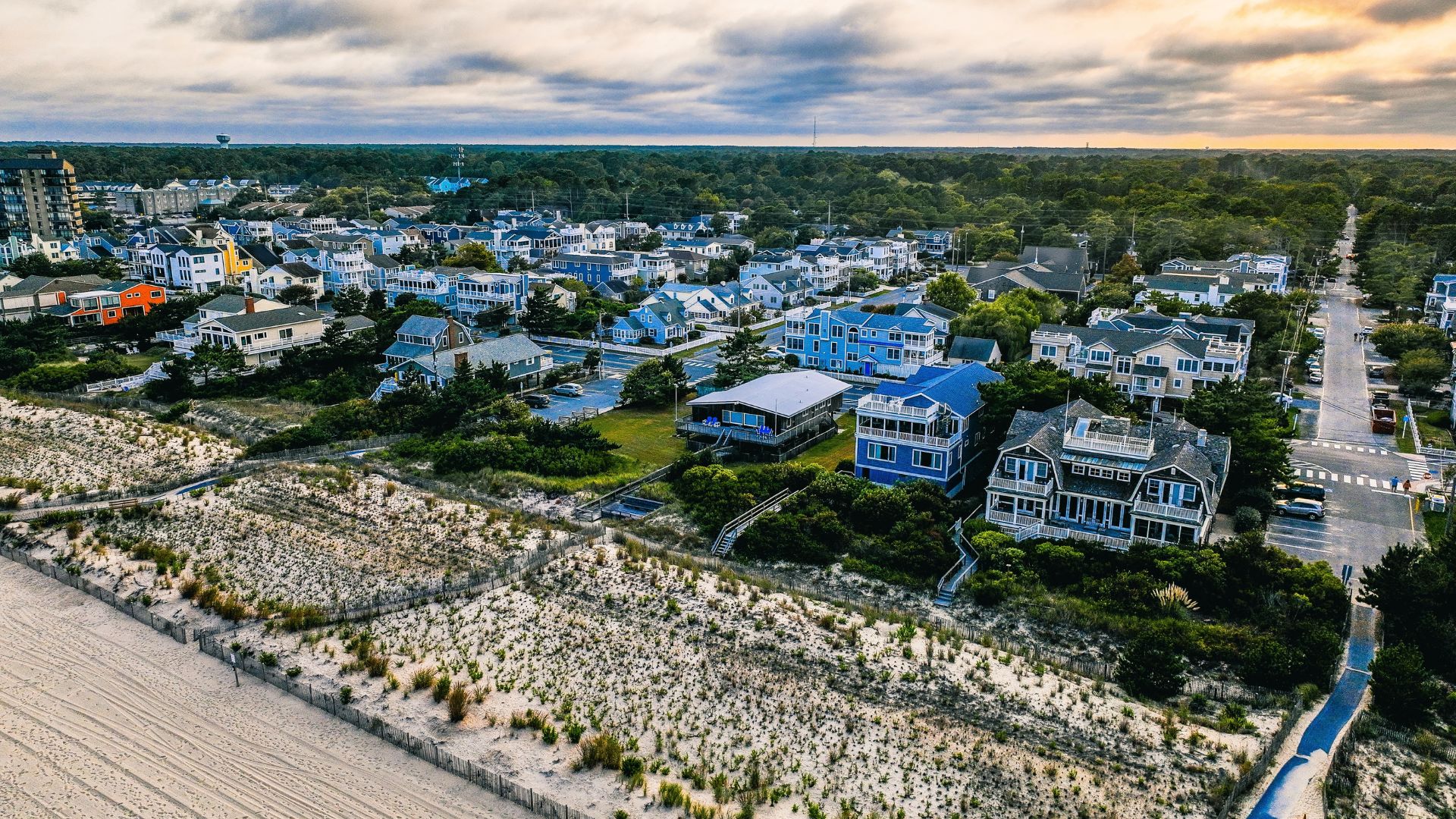 Vista aérea de un pueblo costero con casas coloridas frente a la playa, dunas de arena y vegetación, bajo un cielo parcialmente nublado al atardecer.