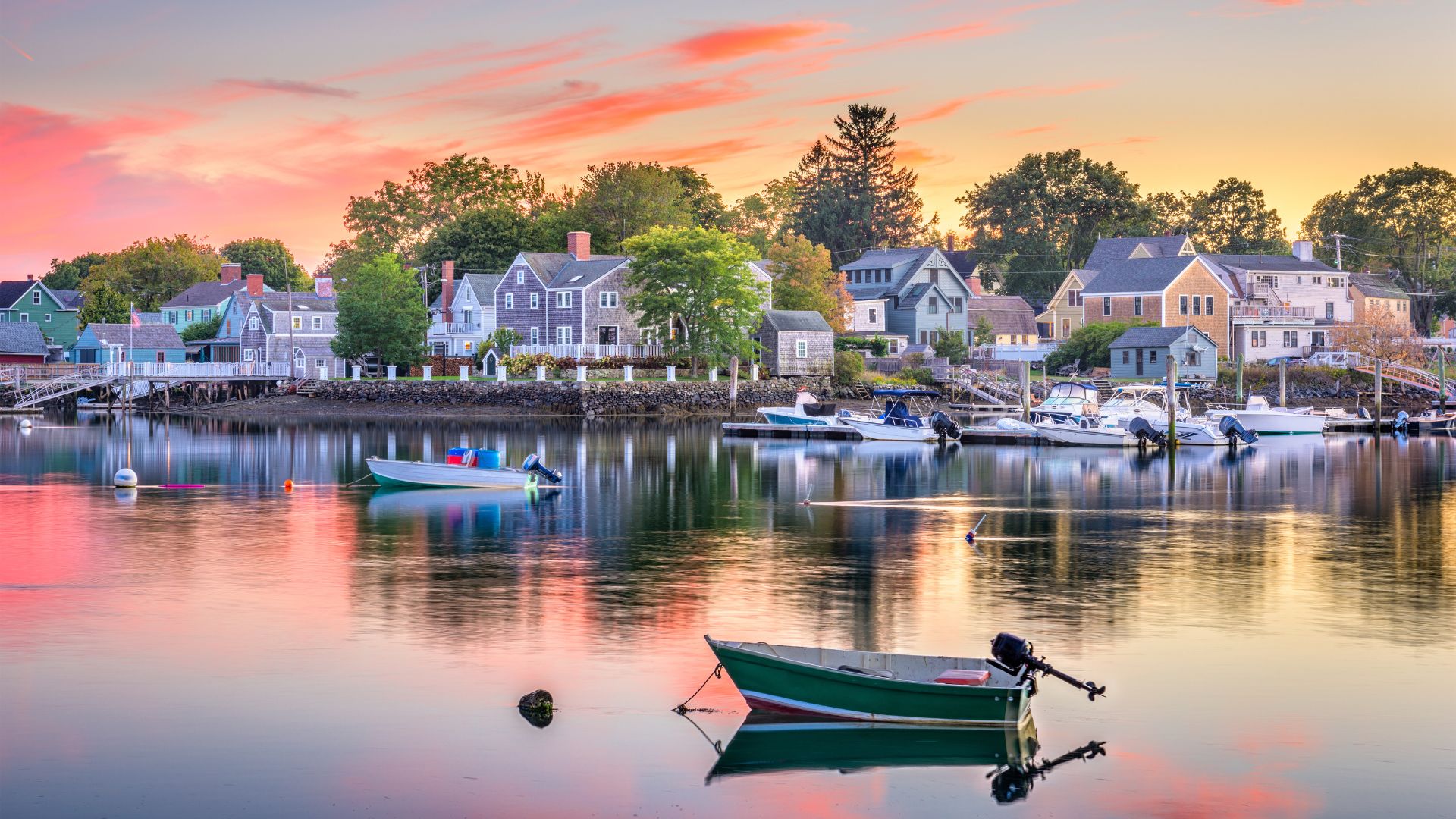 Pequeñas embarcaciones ancladas en un tranquilo puerto frente a casas tradicionales, con reflejos rosados del atardecer sobre el agua.