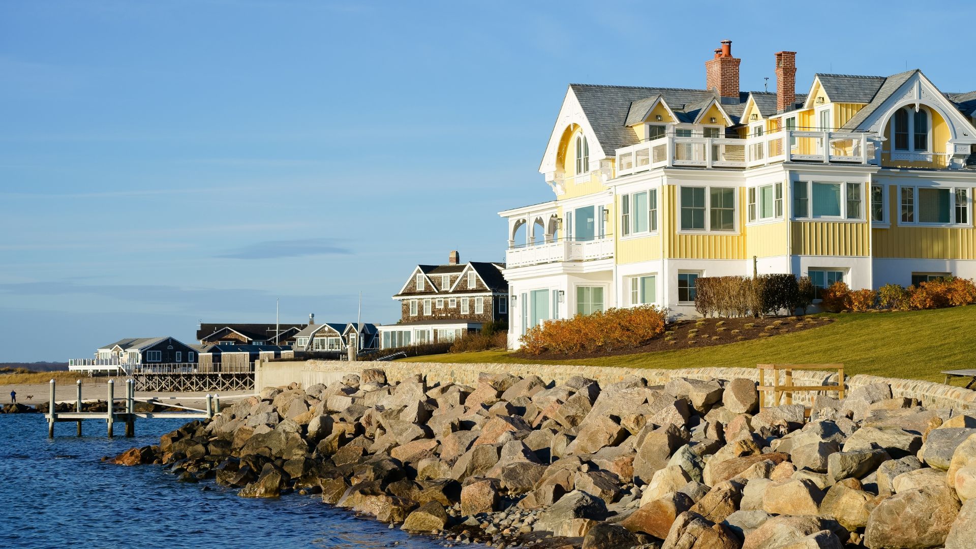 Casa grande de estilo costero pintada en amarillo y blanco sobre un malecón rocoso junto al mar.