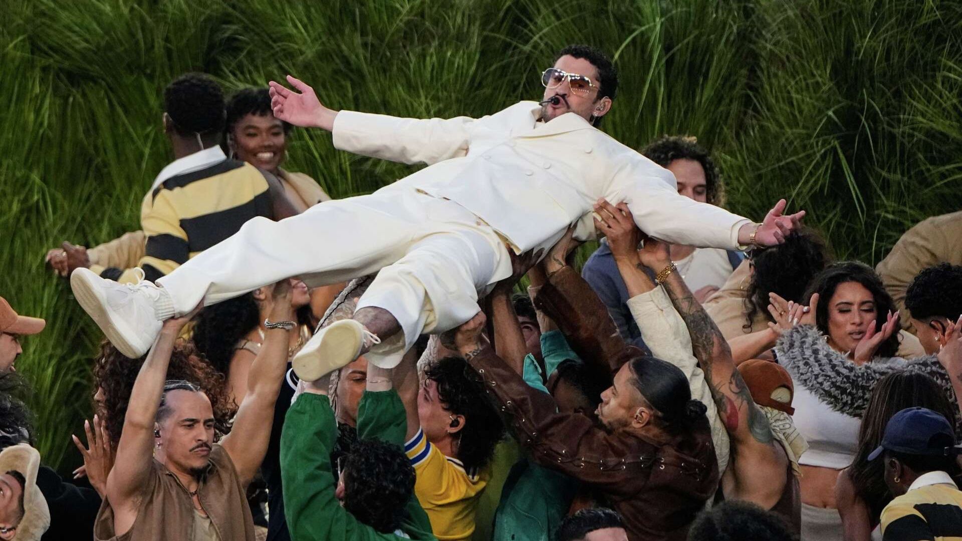 Artista vestido de blanco es alzado por bailarines durante un espectáculo en vivo.