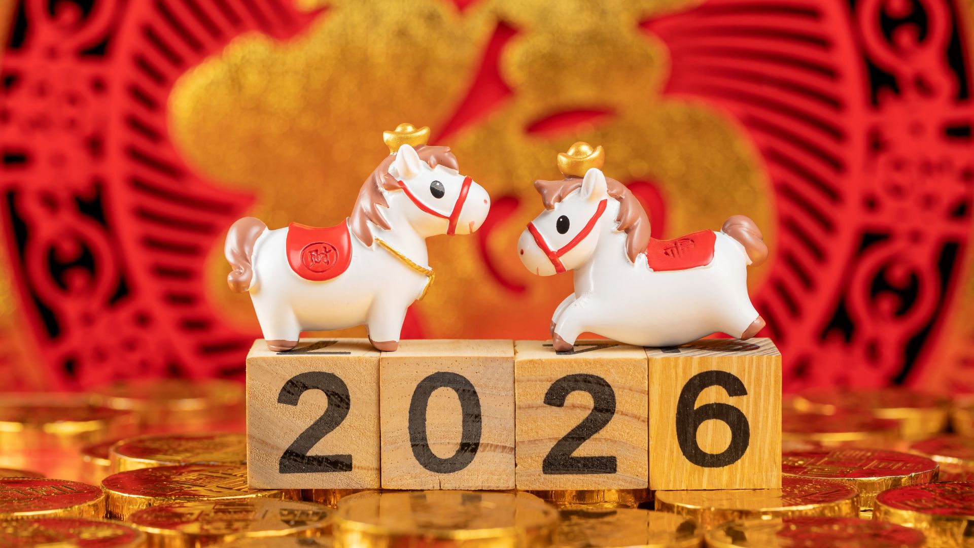 Dos figuras decorativas de caballos blancos sobre bloques de madera con el número 2026, con fondo rojo y monedas doradas.