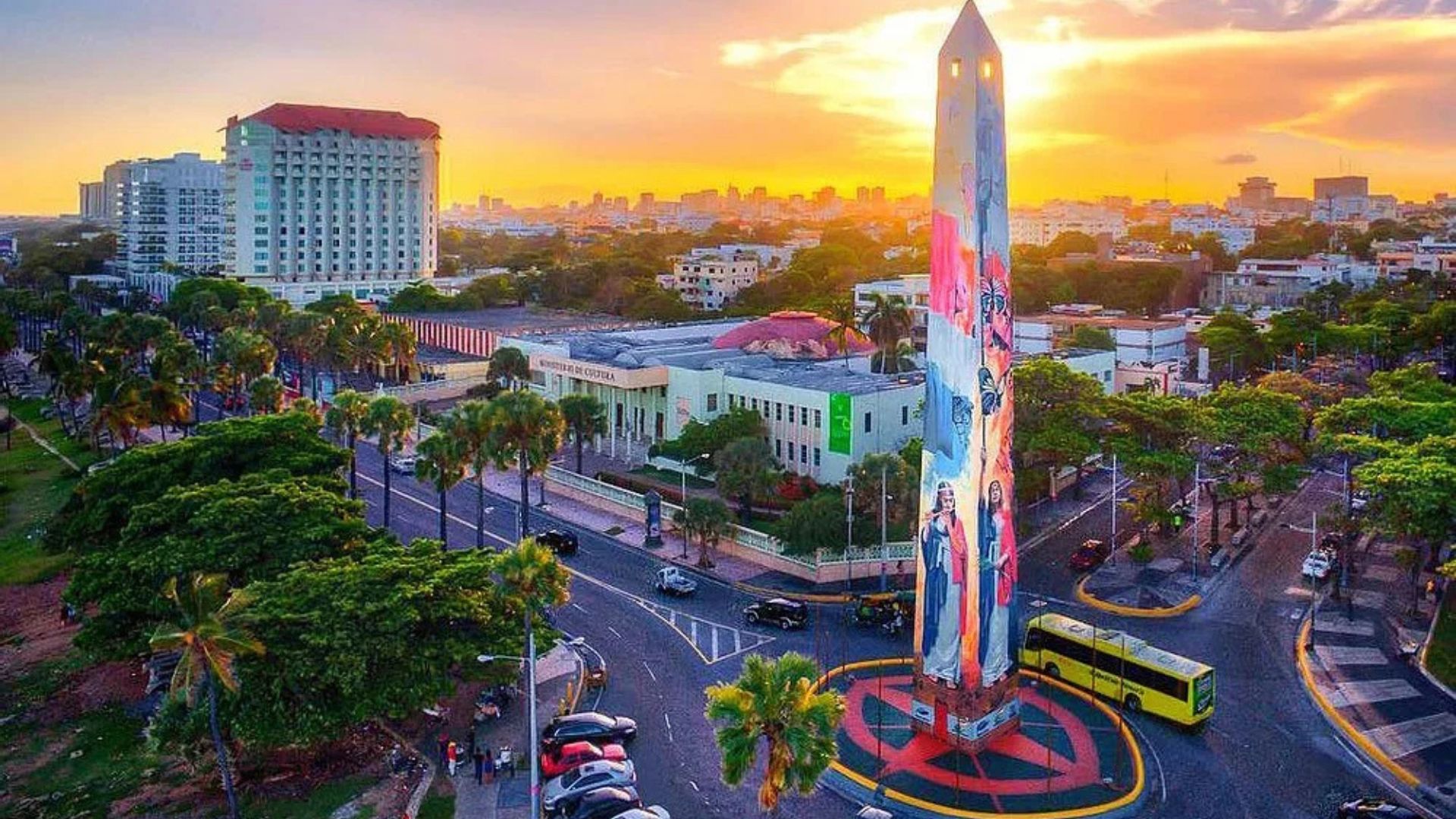 Vista panorámica de la ciudad con un monumento en primer plano y un atardecer en el horizonte.