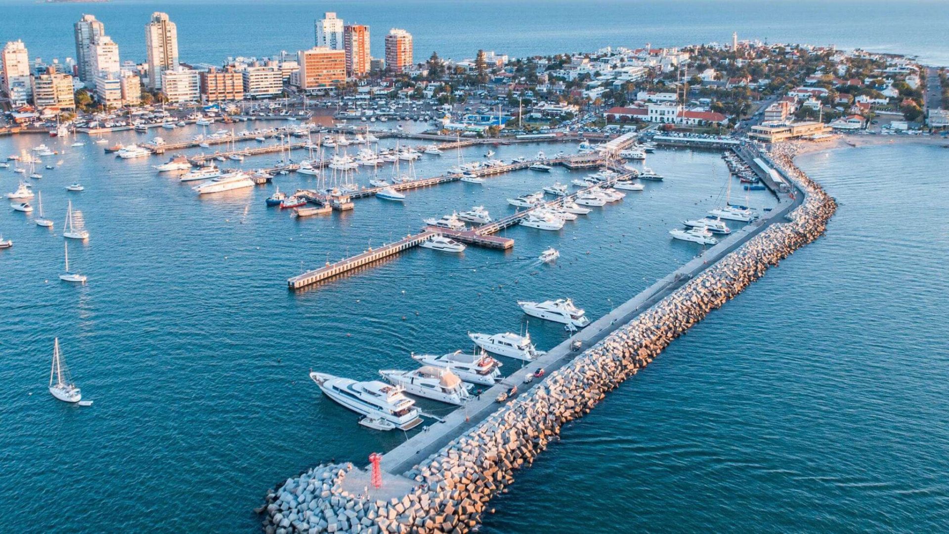 Vista aérea del puerto con yates en el mar, edificios al fondo y el malecón de la ciudad.
