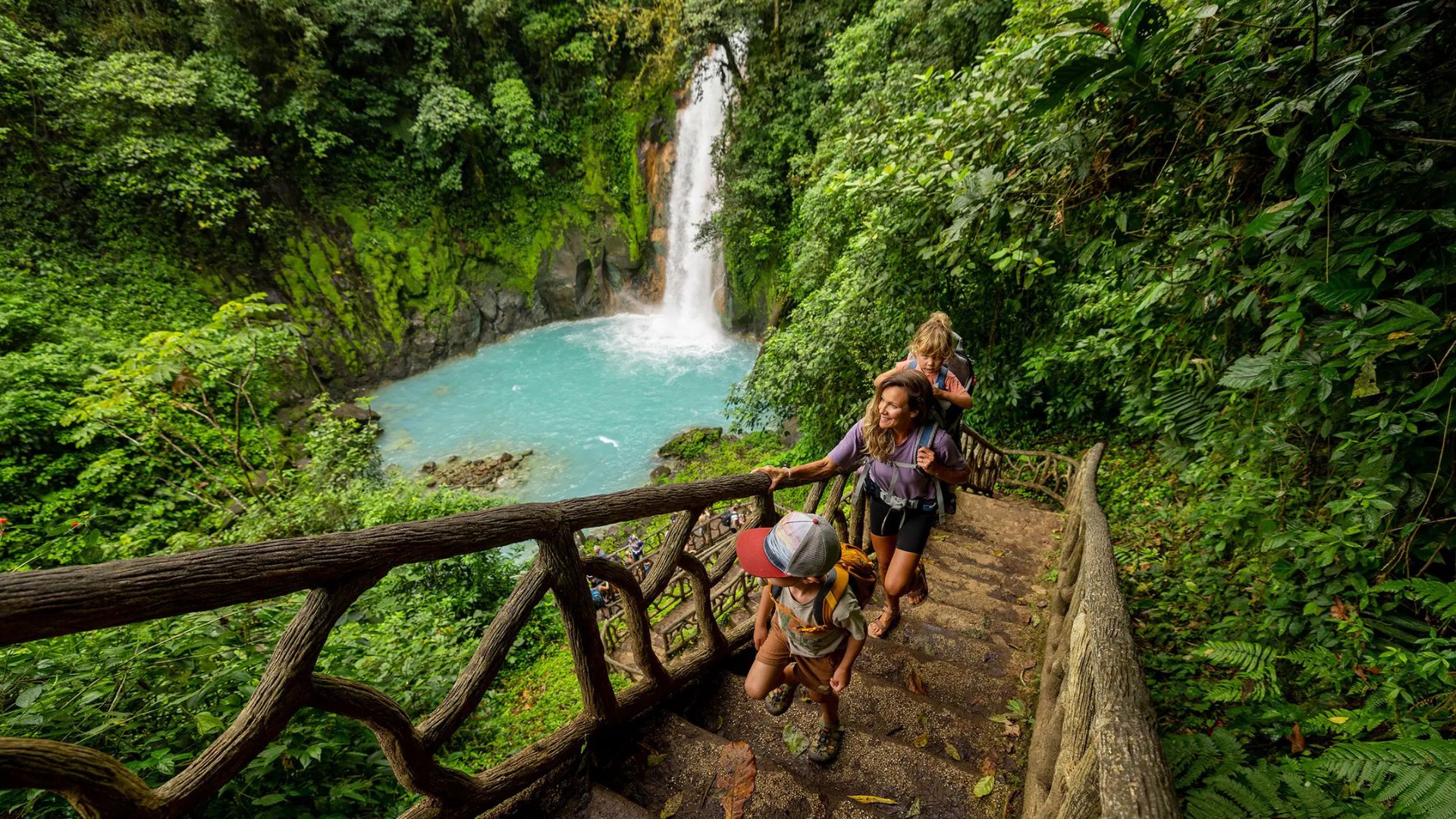 Mujer y dos niños suben por escaleras de madera en la selva hacia una cascada de agua turquesa.