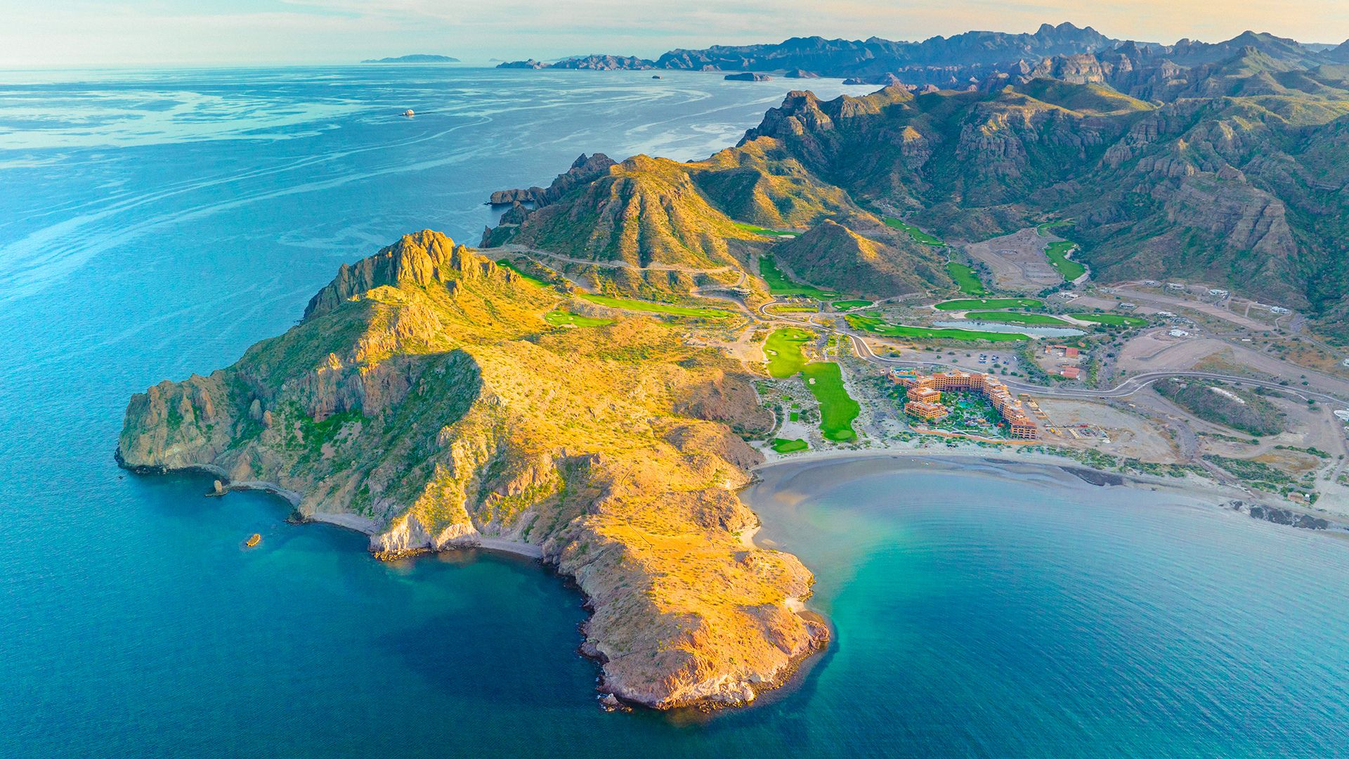 Vista aérea de una península rocosa bañada por aguas turquesas, con montañas áridas y un complejo turístico junto a una bahía.