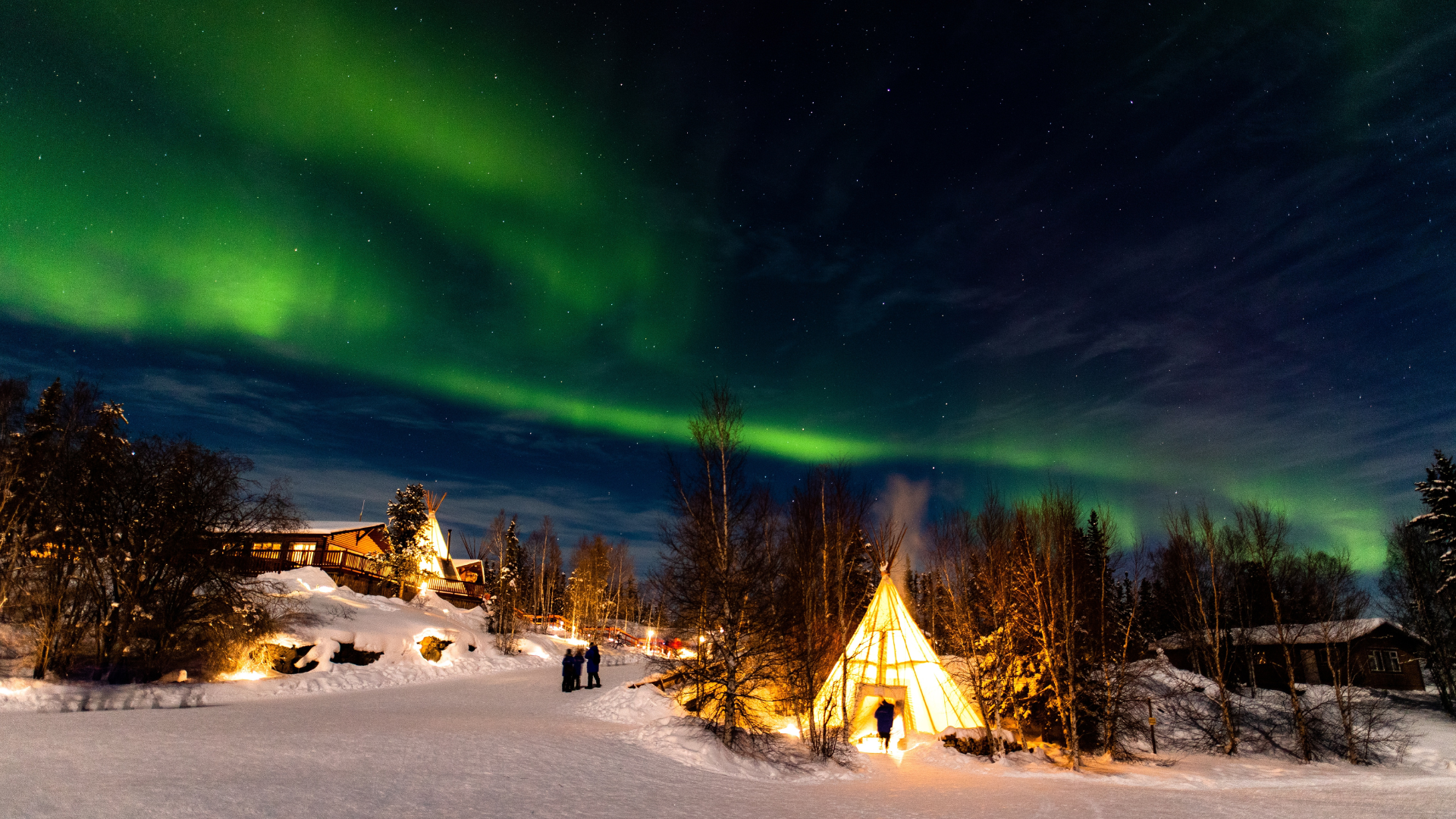 Aurora boreal ilumina el cielo nocturno sobre un paisaje nevado con cabañas y una tienda iluminada en una zona ártica.