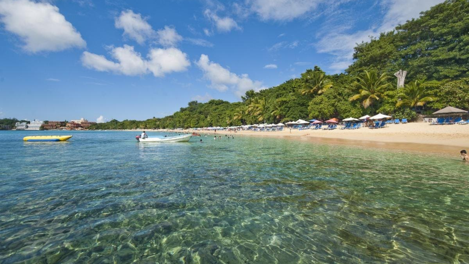 Playa de aguas cristalinas y arena clara en República Dominicana, con botes, sombrillas y vegetación tropical al fondo en un día soleado.