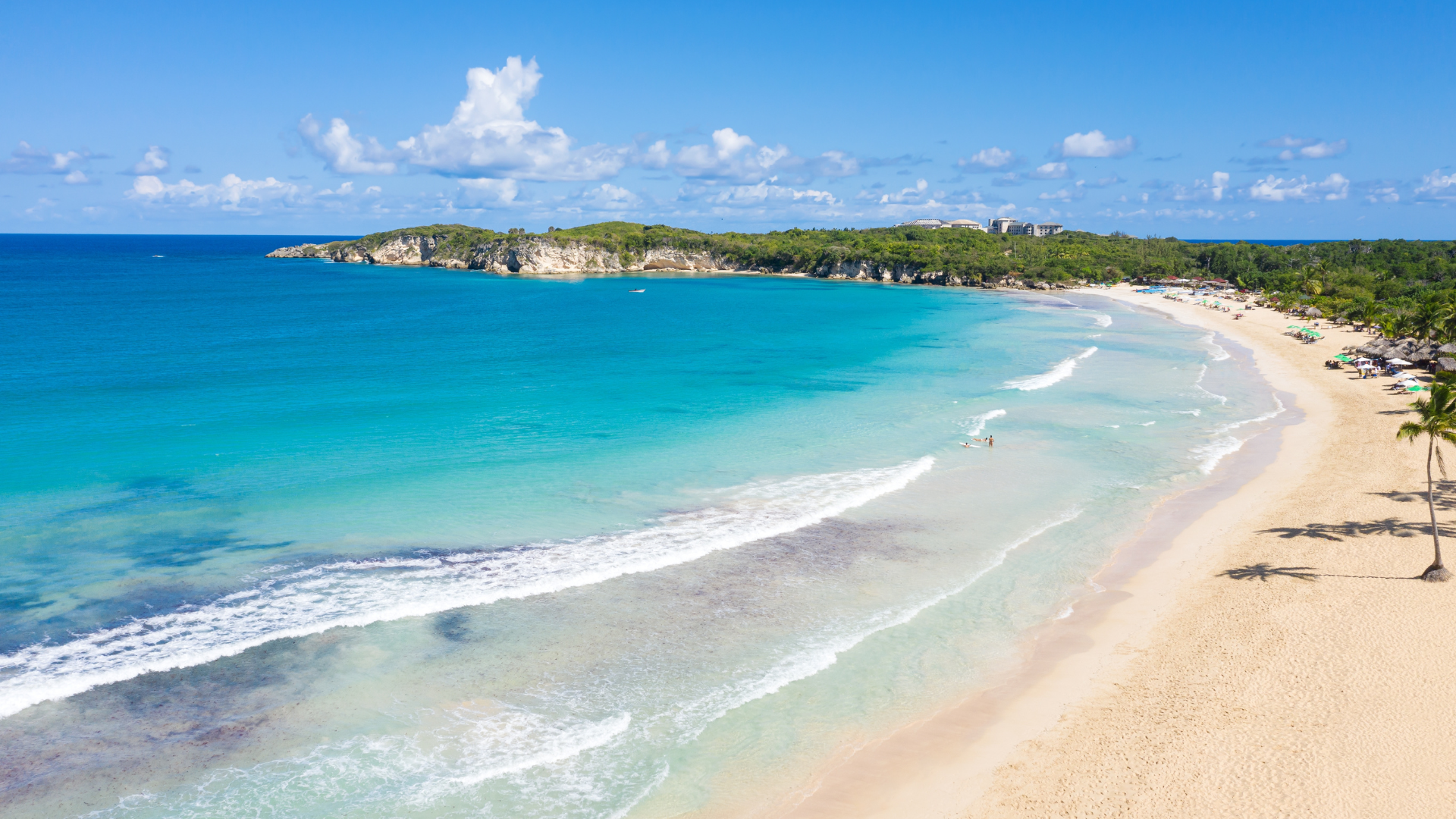 Vista panorámica de una playa de aguas turquesas y arena blanca en República Dominicana, con palmeras, oleaje suave y colinas verdes al fondo.