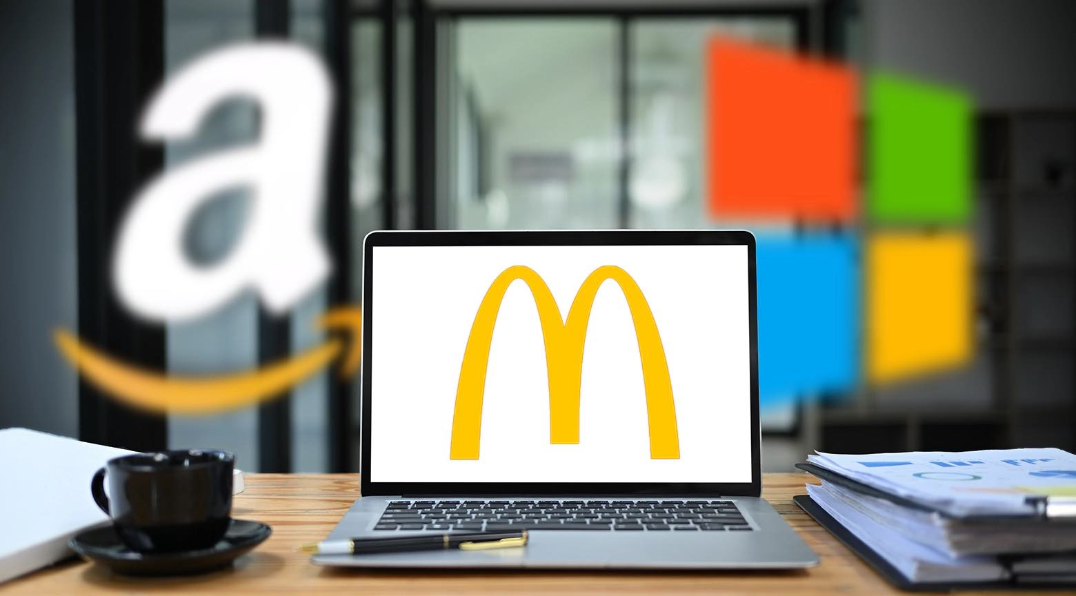 Un portátil con el logo de McDonald's en la pantalla, rodeado de los logotipos borrosos de Amazon y Microsoft, con una taza de café sobre una mesa de trabajo.