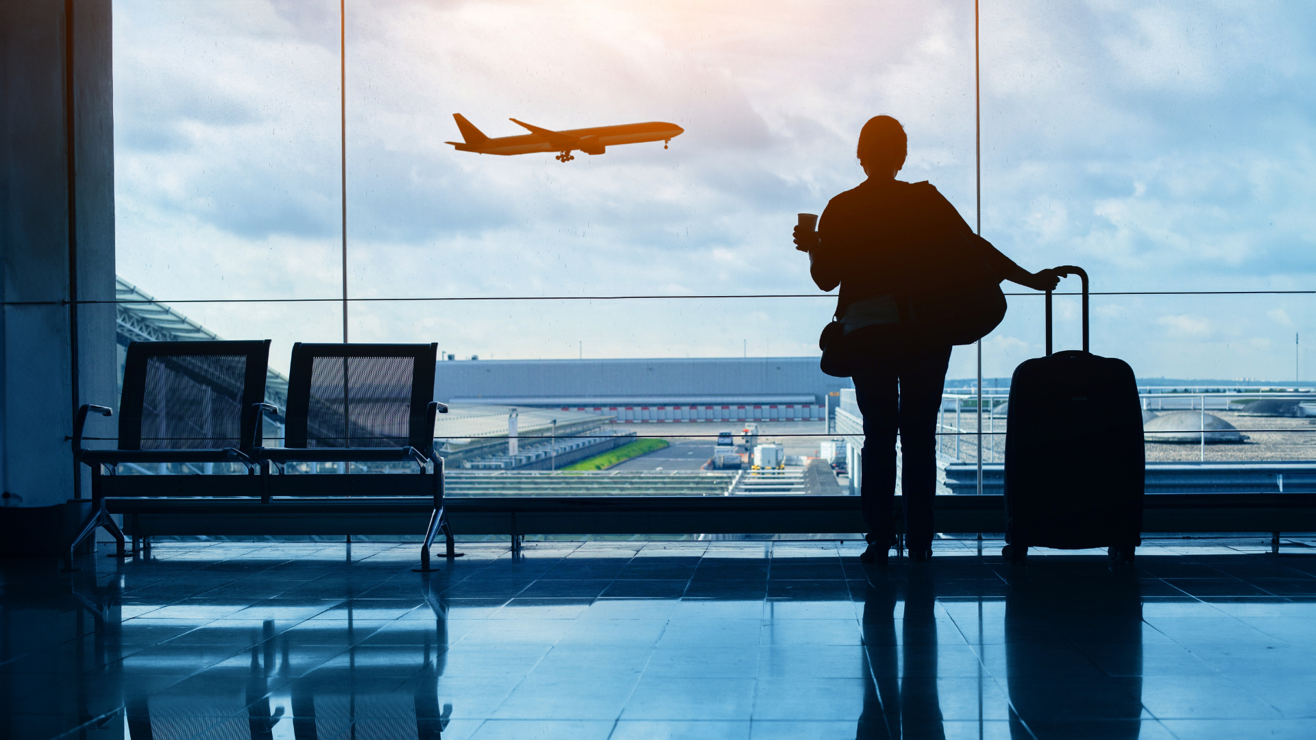Persona con maleta observa un avión despegar desde la ventana de un aeropuerto.