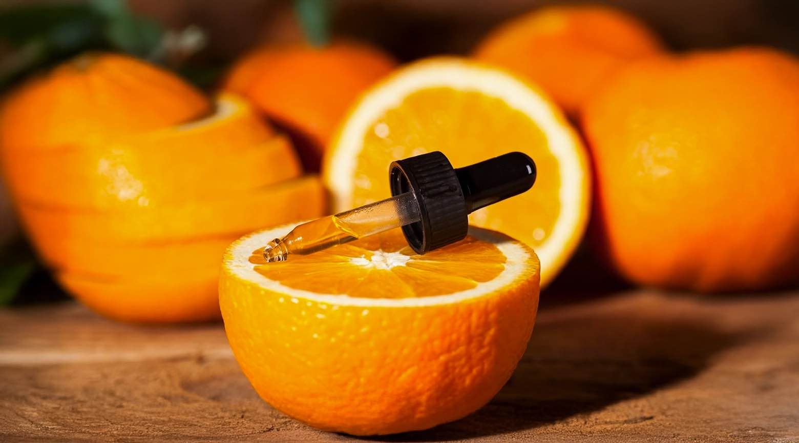 Mitad de una naranja sobre una mesa de madera con un gotero que contiene líquido, rodeada de otras naranjas.