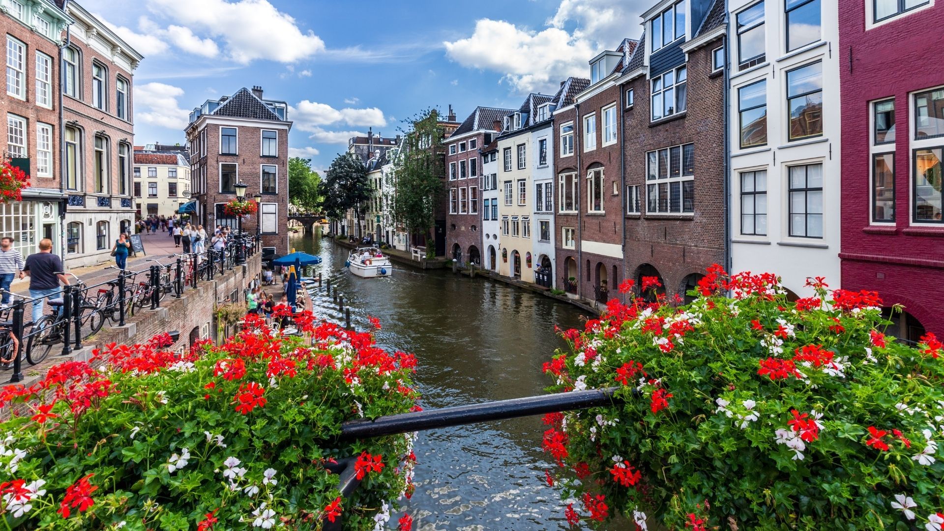 Canal en una ciudad europea rodeado de edificios antiguos, con flores rojas y blancas en primer plano.
