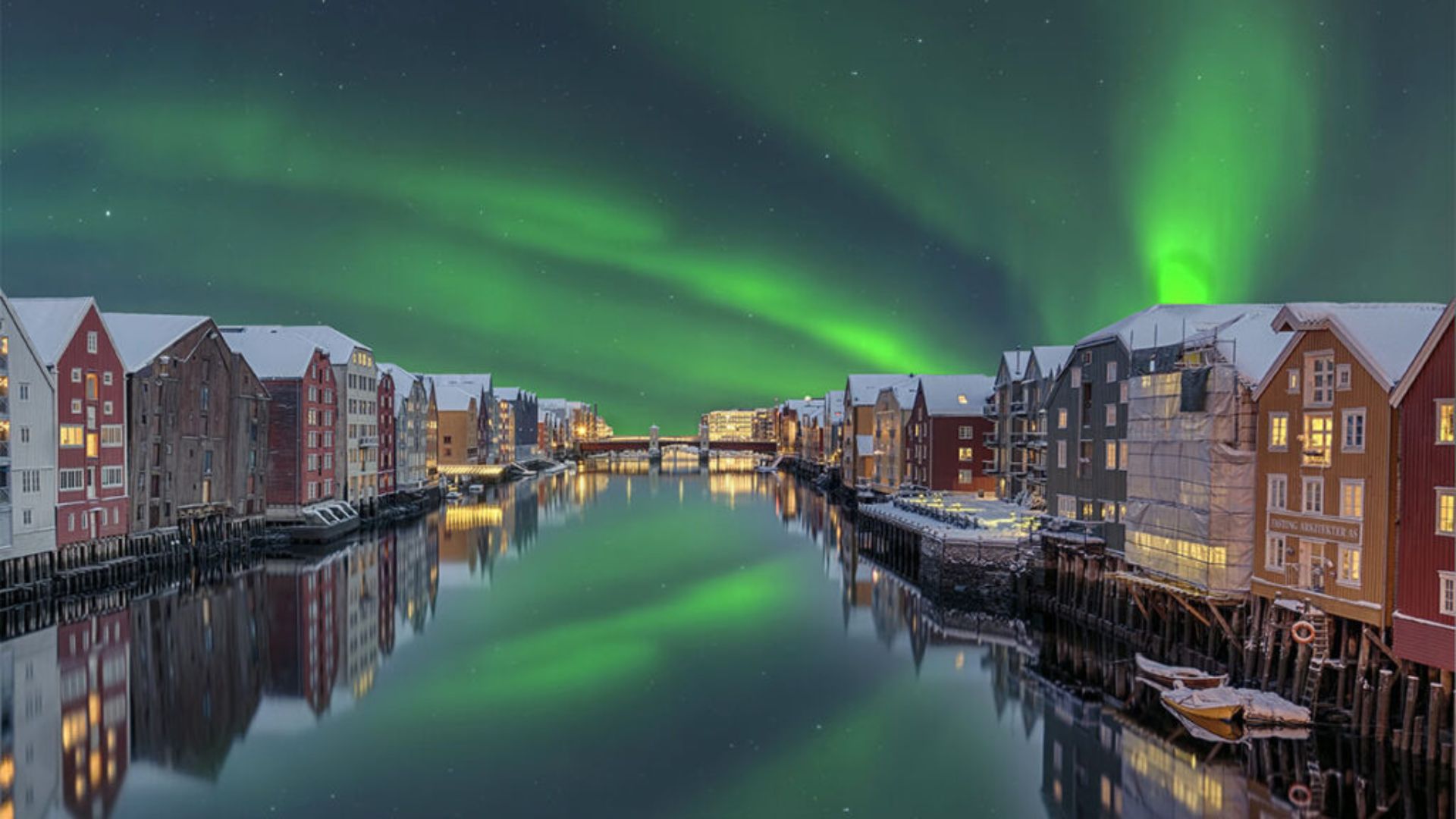 Vista nocturna de una ciudad con canales en Noruega bajo las auroras boreales, con edificios reflejados en el agua.