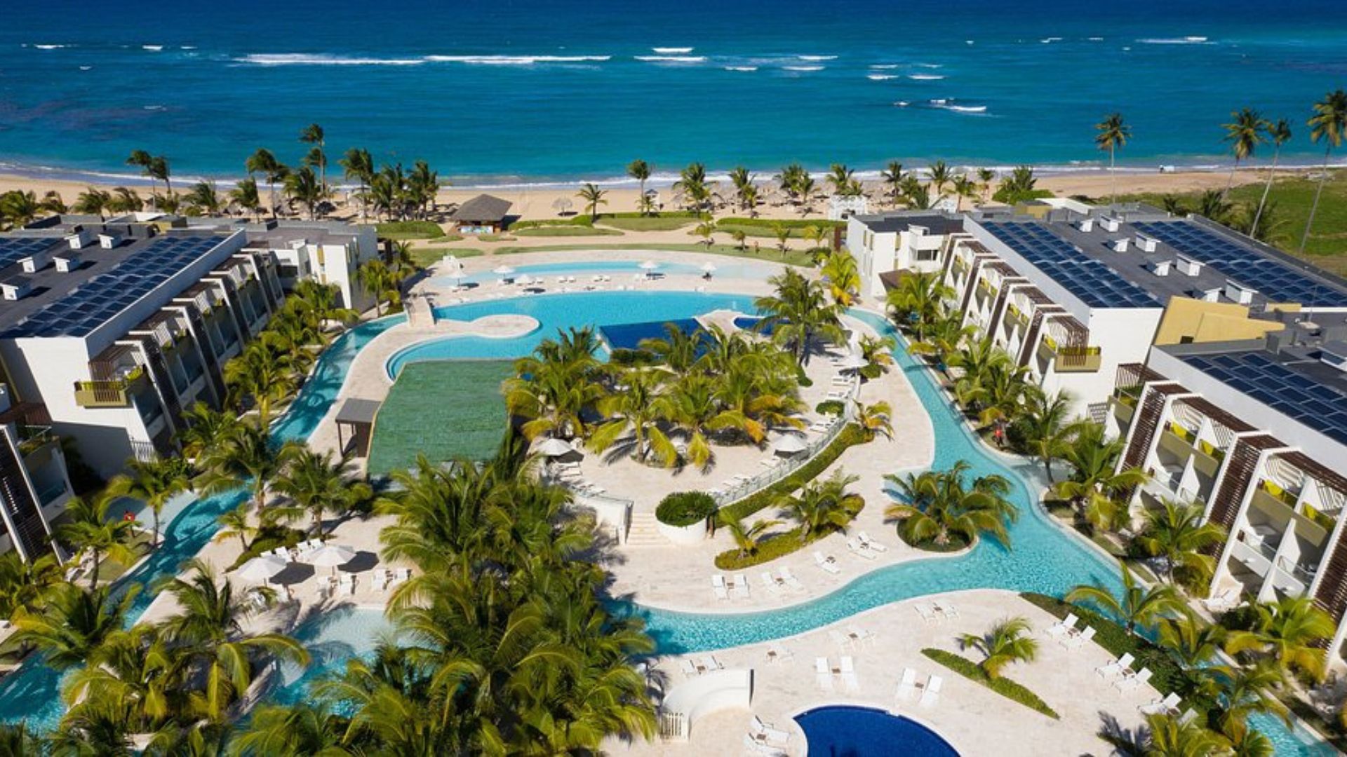 Vista aérea de un complejo hotelero con piscinas rodeadas de palmas, frente al mar en Punta Cana.