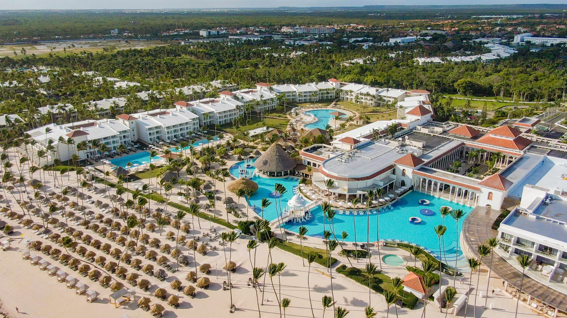 Vista aérea de un resort frente a la playa en Punta Cana, con áreas de piscinas y palapas en la arena.