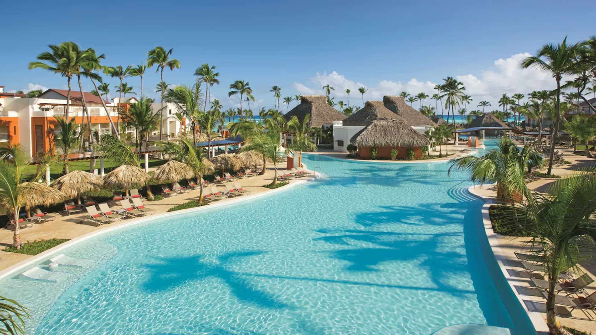 Vista de una piscina en un resort en Punta Cana, rodeada de sombrillas y palmeras, con el mar en el fondo.
