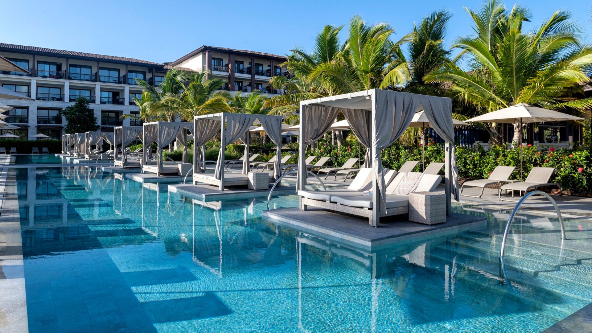 Vista de una piscina con camas balinesas y sombrillas, rodeada de palmeras y edificios de resort.