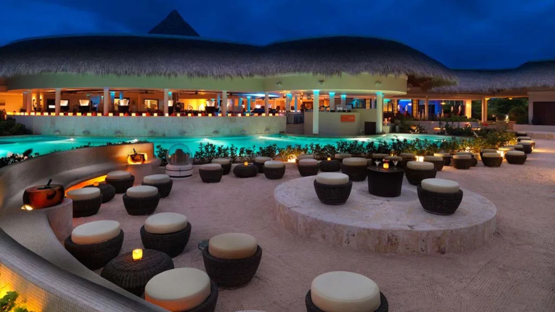 Restaurante y área lounge de un resort de lujo en Punta Cana con piscina iluminada y techo de palma.