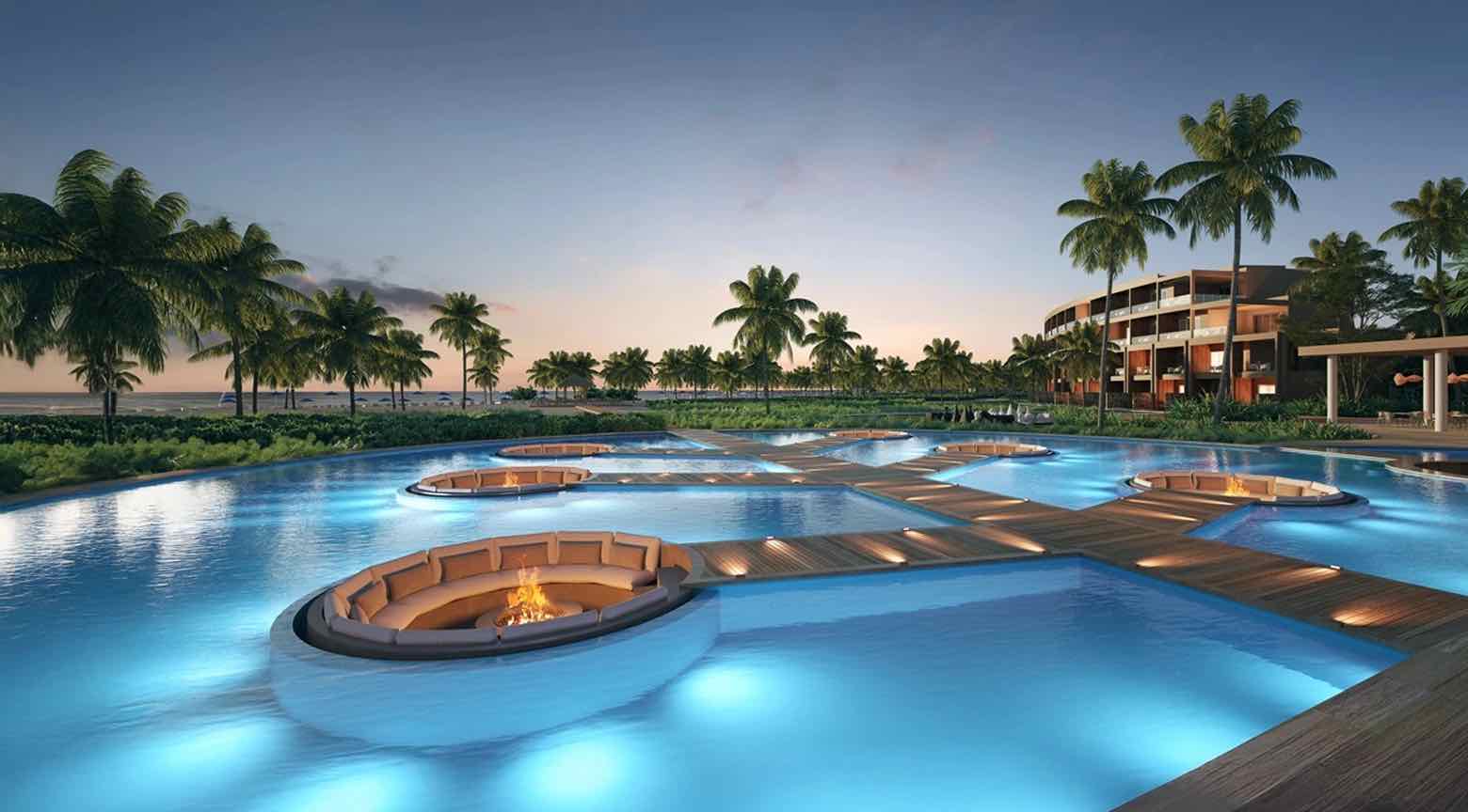 Resort de lujo con piscina iluminada, áreas de descanso circulares y palmeras junto al mar al atardecer.